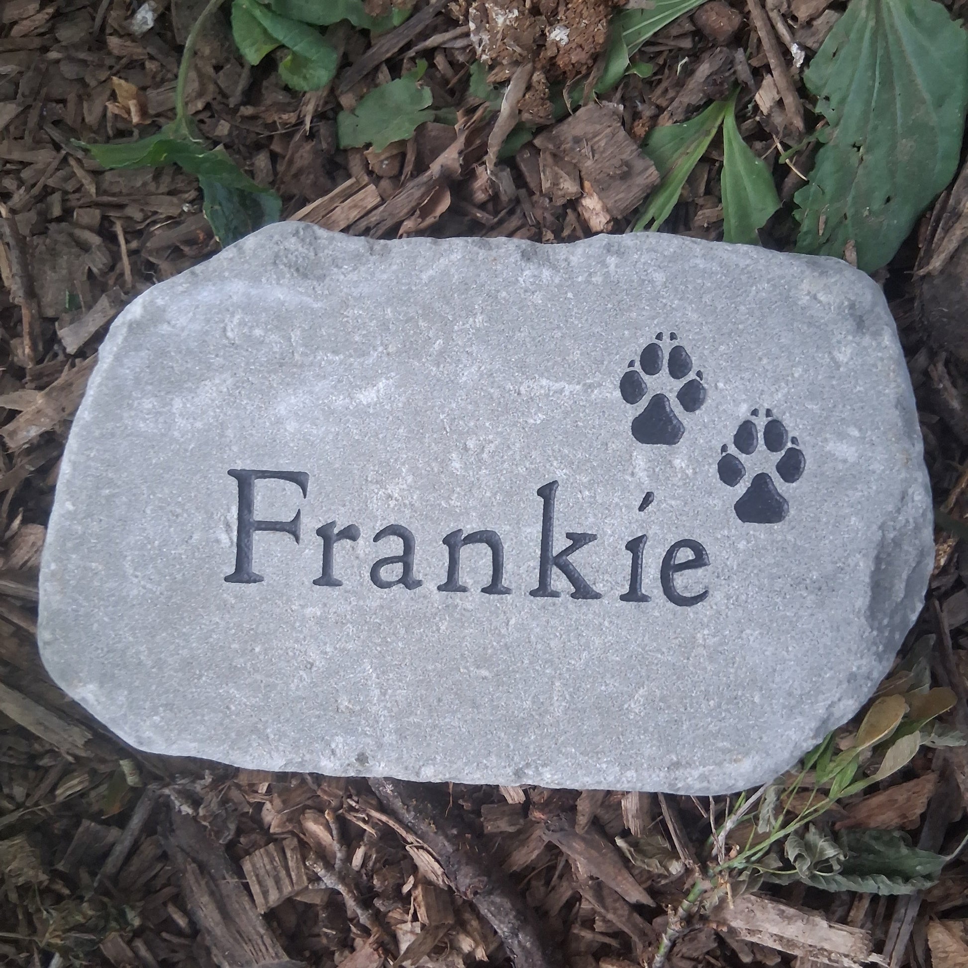 A personalized gray pet memorial garden stone engraved with the name 'Frankie' and two paw prints, placed on a bed of brown wood chips and green leaves.
