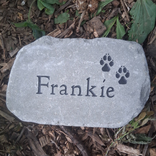 A personalized gray pet memorial garden stone engraved with the name 'Frankie' and two paw prints, placed on a bed of brown wood chips and green leaves.