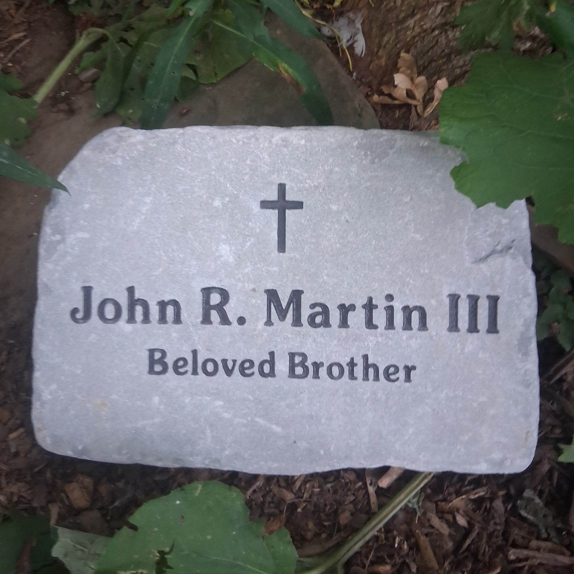 A personalized gray stone grave marker with a cross symbol and the engraved text ' Beloved Brother', placed outdoors on the ground among green leaves and mulch.