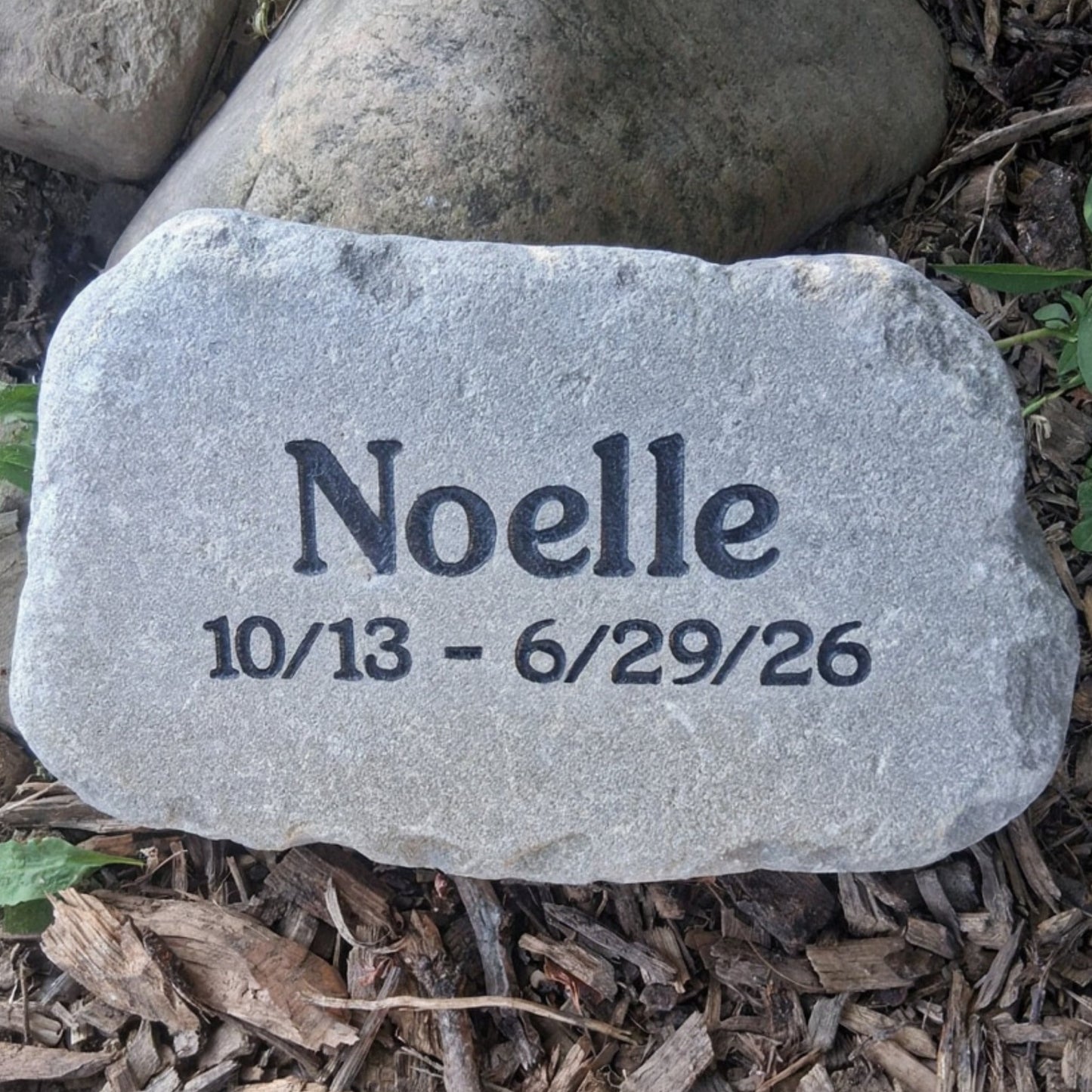 A personalized gray memorial garden stone engraved with the name 'Noelle' and the dates '10/13 - 6/29/26', placed on a bed of brown wood chips and mulch outdoors.