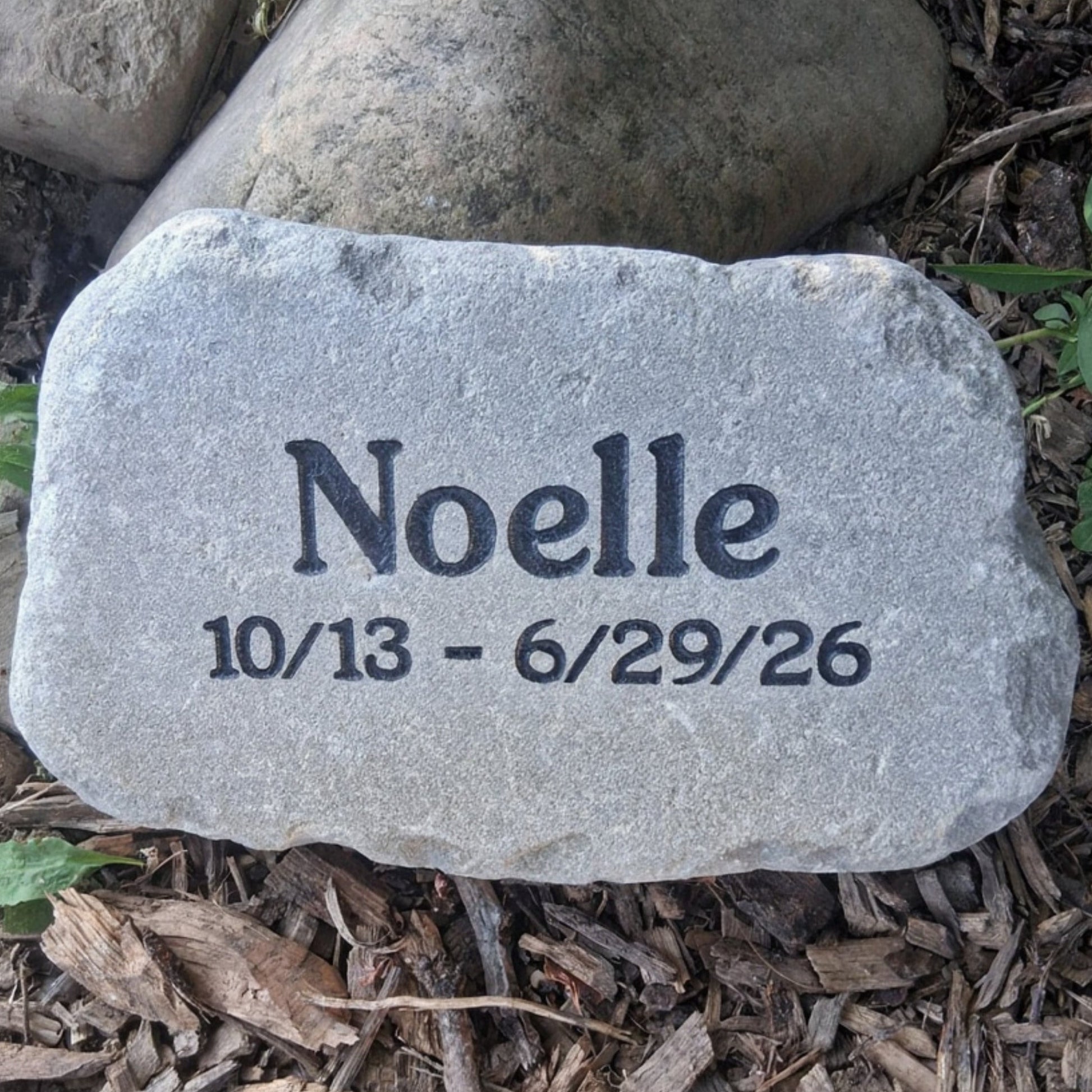 A personalized gray memorial garden stone engraved with the name 'Noelle' and the dates '10/13 - 6/29/26', placed on a bed of brown wood chips and mulch outdoors.