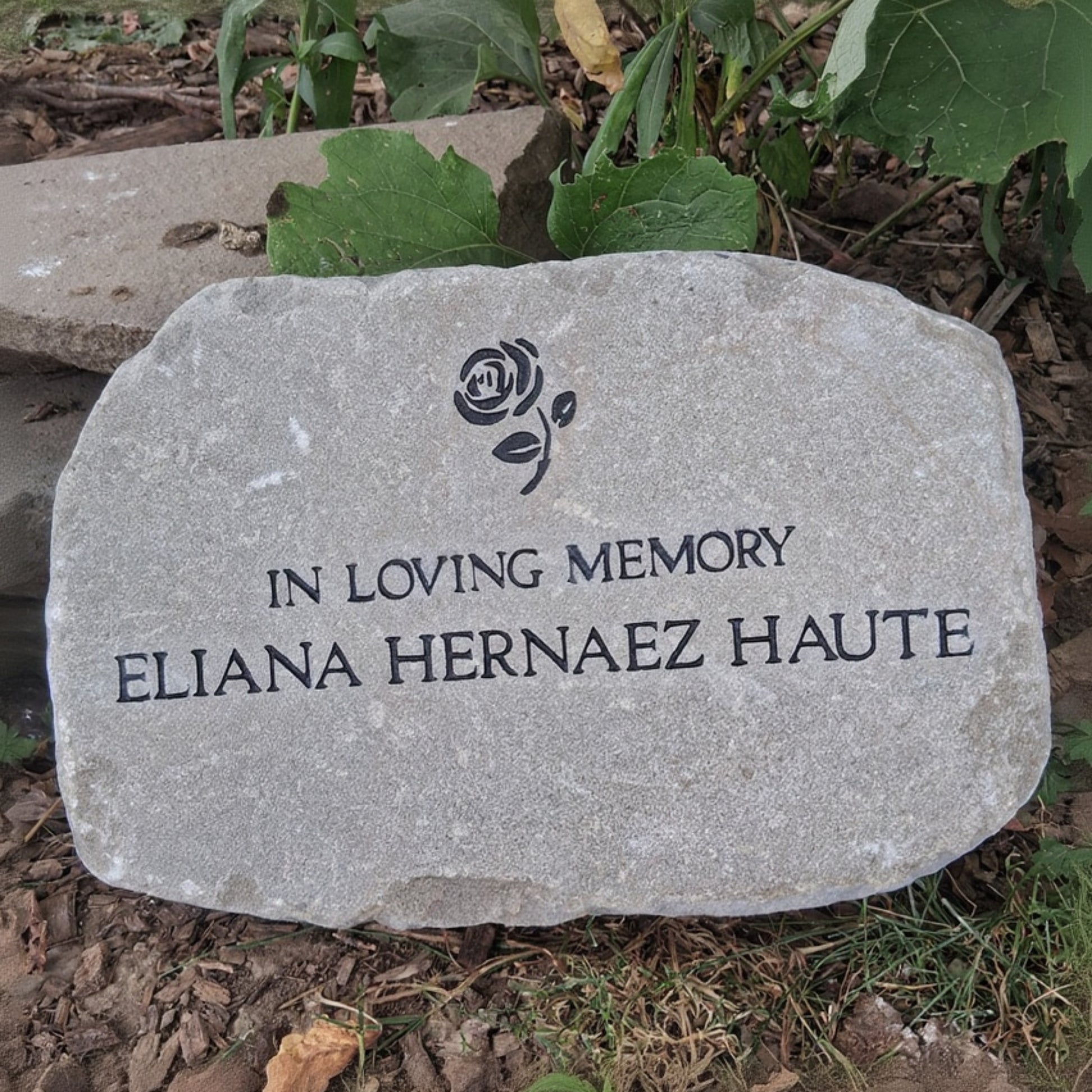 A custom engraved memorial garden stone with a rose icon and the text 'IN LOVING MEMORY ELIANA HERNAEZ HAUTE' placed outdoors on mulch and grass.