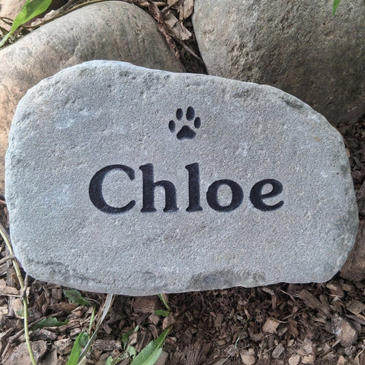A personalized gray garden stone engraved with the name 'Chloe' and a small paw print, placed on a bed of mulch and green plants.
