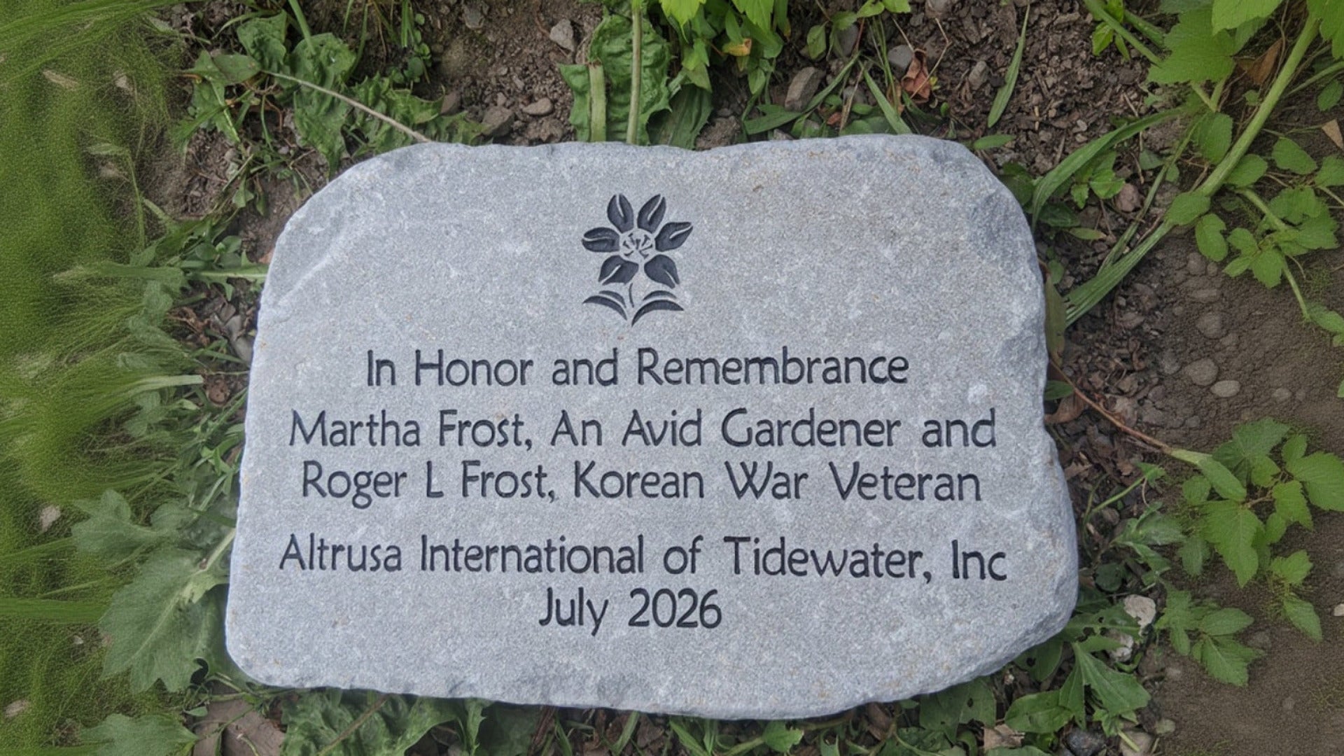 A rectangular gray memorial stone with engraved black text and a small flower emblem, placed outdoors on grass and soil.