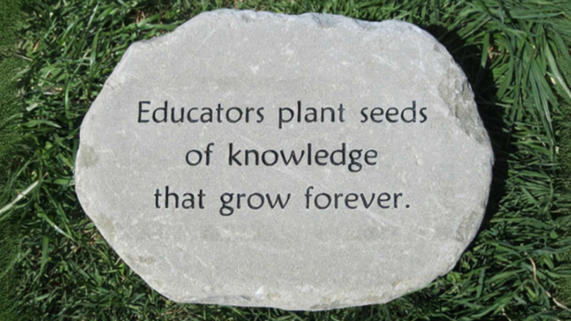 A round brown stone plaque engraved with the quote 'Educators plant seeds of knowledge that grow forever.' placed on a green grass background.