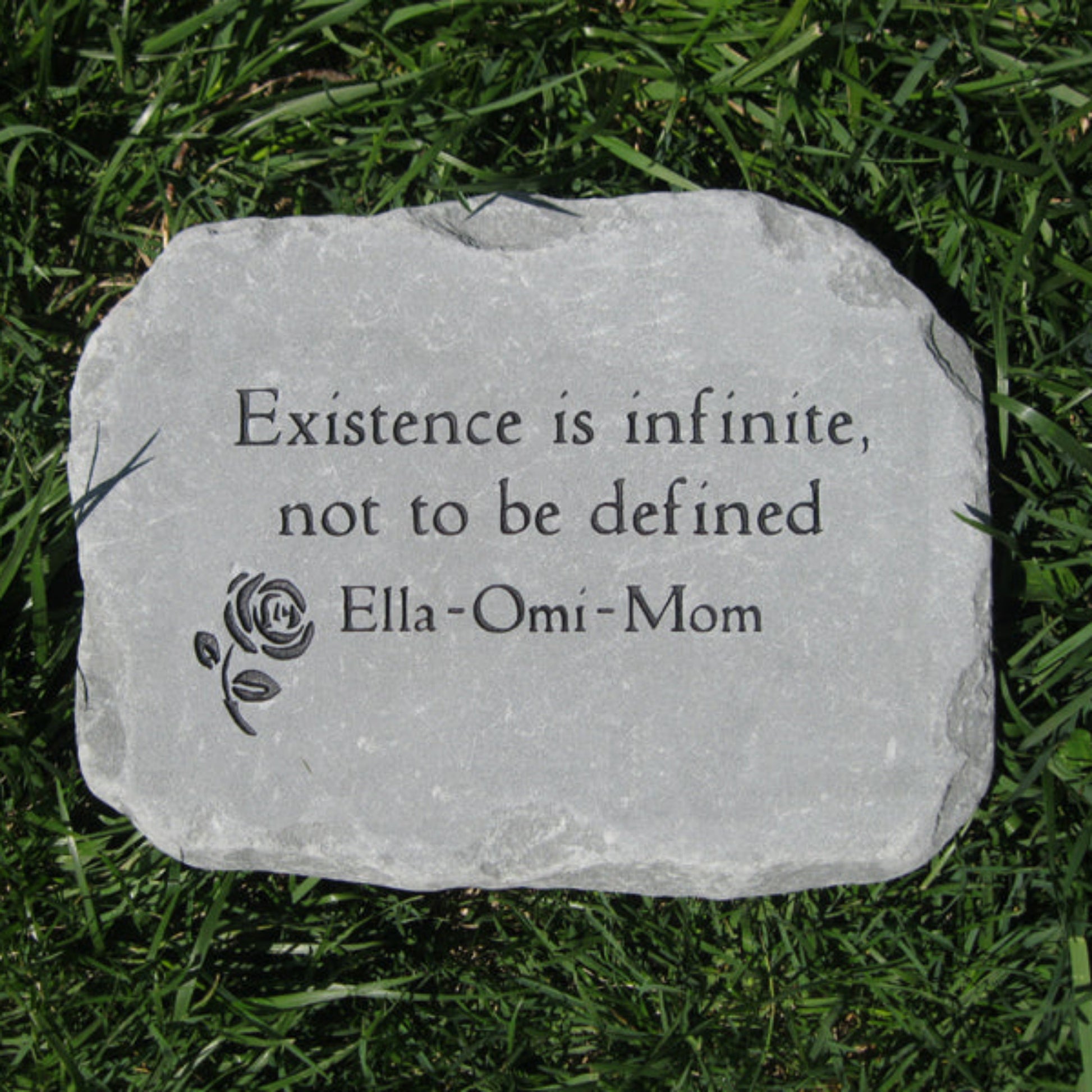 A custom engraved  stone memorial plaque lying on green grass, featuring the quote 'Existence is infinite, not to be defined' and the name 'Ella-Omi-Mom' alongside a small rose engraving.