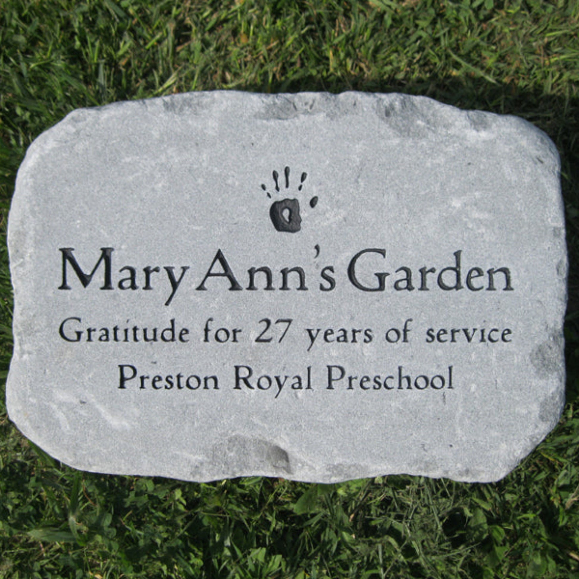 A rectangular light gray memorial stone with engraved black text reading 'Mary Ann's Garden' and 'Gratitude for 27 years of service Preston Royal Preschool,' placed on green grass.