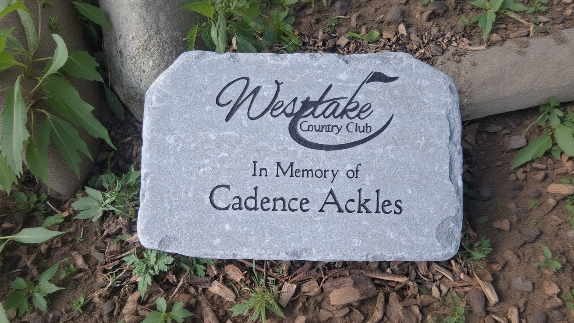 A personalized gray memorial garden stone with black engraved lettering and customized with custom logo placed on a bed of mulch and small green plants.