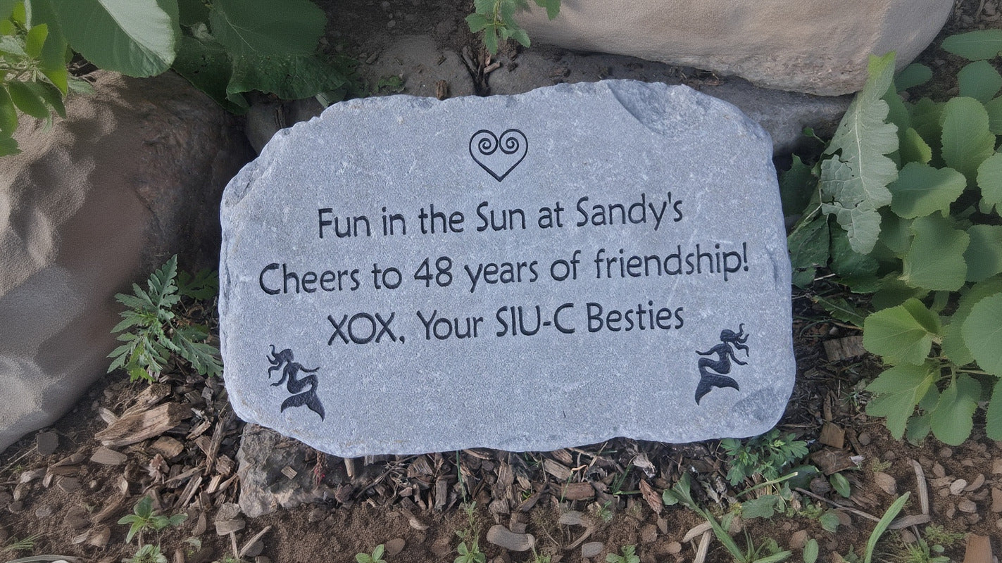 A custom engraved gray stone plaque placed outdoors on mulch, featuring the text 'Fun in the Sun at Sandy's Cheers to 48 years of friendship! XOX, Your SIU-C Besties' along with a heart logo and mermaid designs.