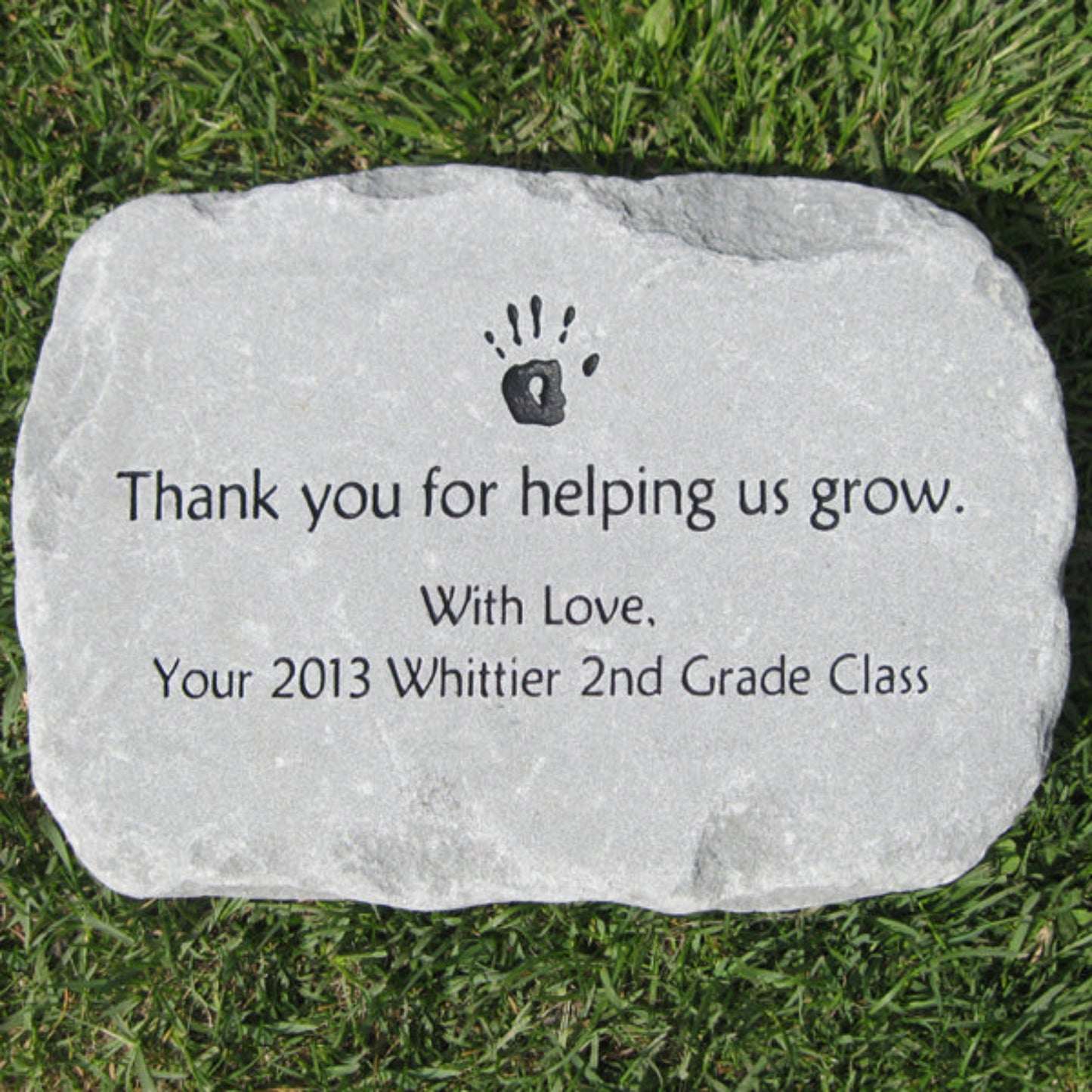 A personalized light gray teacher class gift garden stone engraved with a black hand print and the text 'Thank you for helping us grow. With Love, Your 2013 Whittier 2nd Grade Class', placed on green grass.