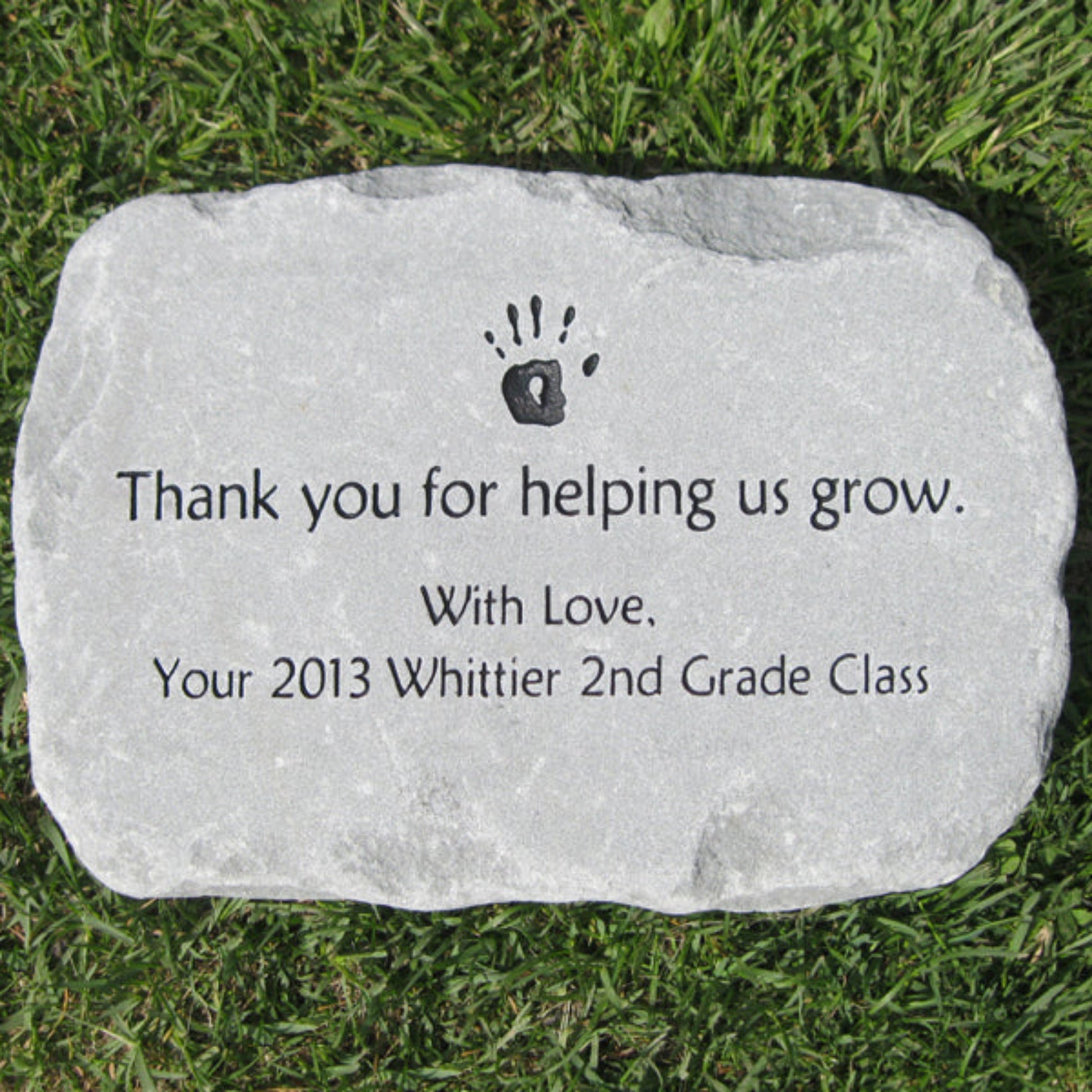 A personalized light gray teacher class gift garden stone engraved with a black hand print and the text 'Thank you for helping us grow. With Love, Your 2013 Whittier 2nd Grade Class', placed on green grass.