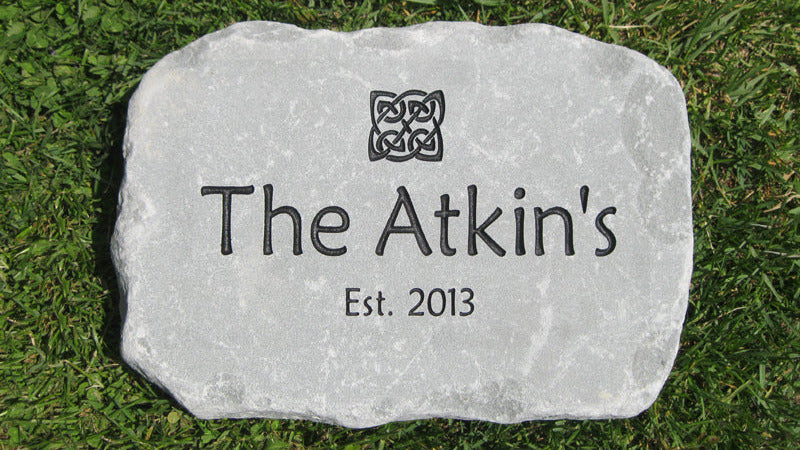 A personalized light gray stone address marker engraved with 'The Atkin's Est. 2013' and a Celtic knot design, placed on green grass.