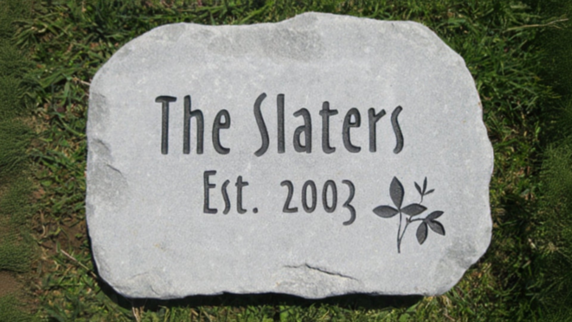 A personalized natural stone address marker engraved with 'The Slaters Est. 2003' and a small leaf design, placed on green grass.