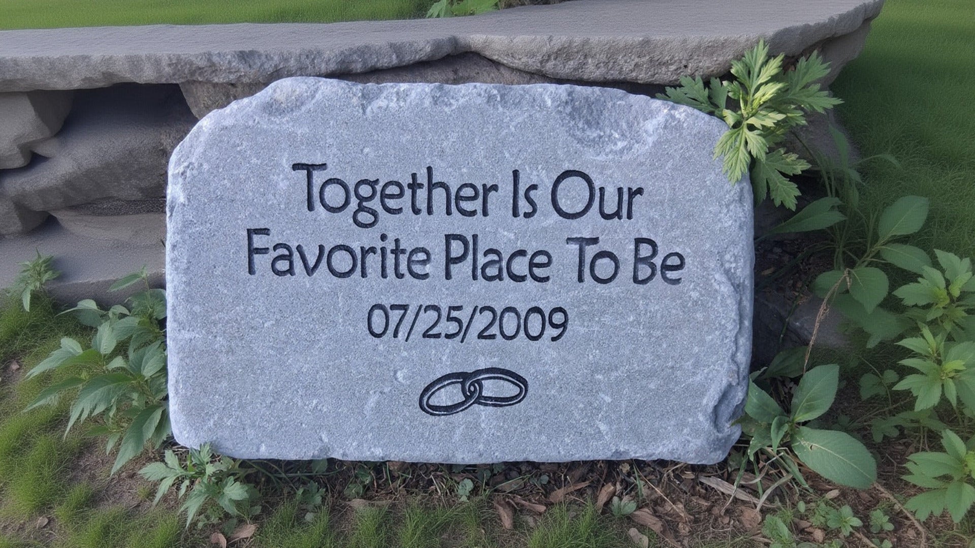 A custom engraved bluestone anniversary stone placed outdoors on a bed of mulch and small green plants. The stone features the engraved text 'Together Is Our Favorite Place To Be 07/25/2009' alongside a simple black wedding ring image