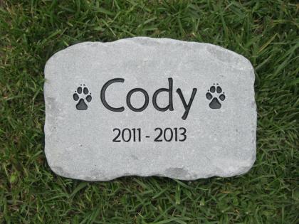 A personalized pet memorial stone engraved with the name 'Cody', paw prints, and the years '2011 - 2013', placed on green grass.