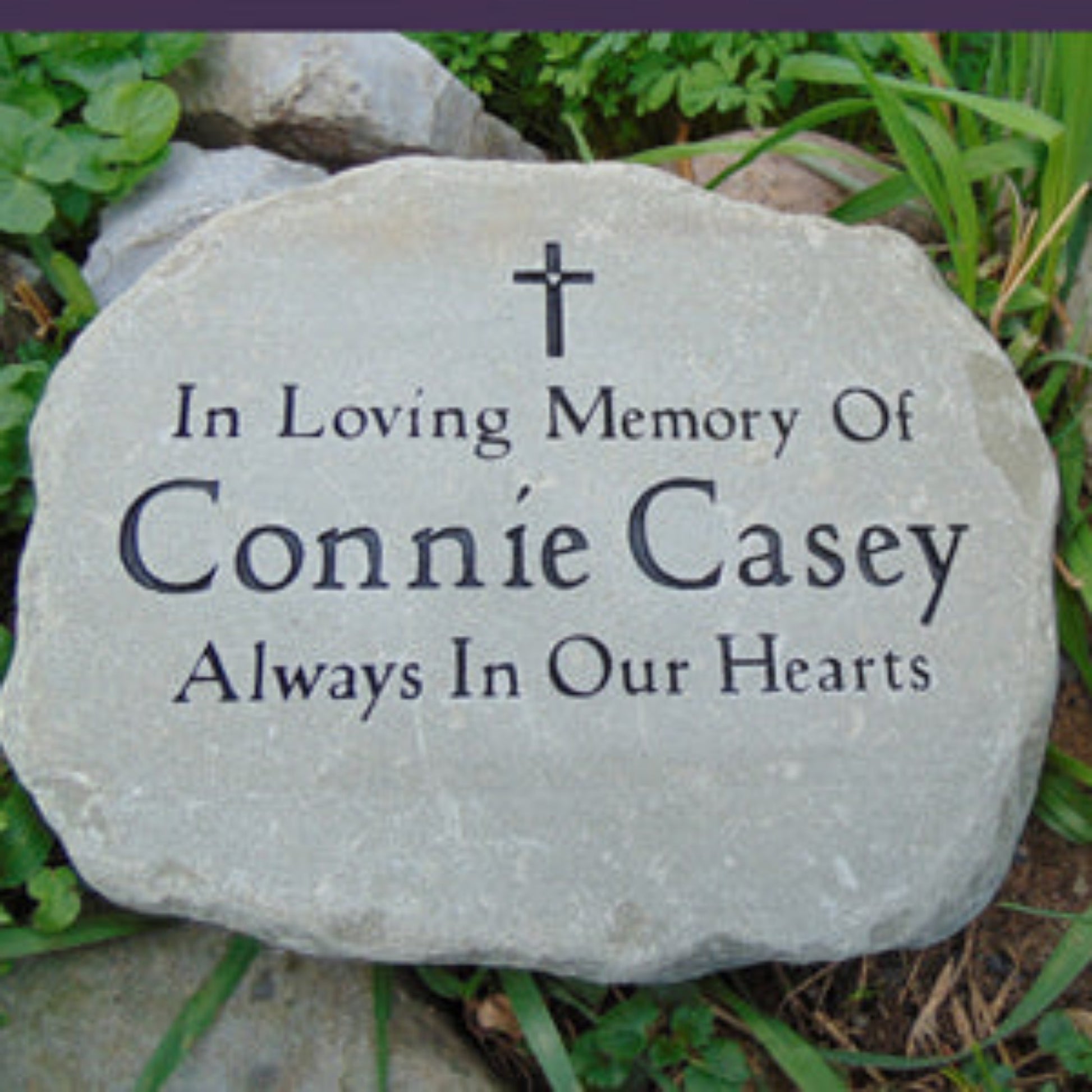 A personalized memorial garden stone with a cross engraved, reading 'In Loving Memory Of and Always In Our Hearts', placed outdoors among green grass and other rocks.