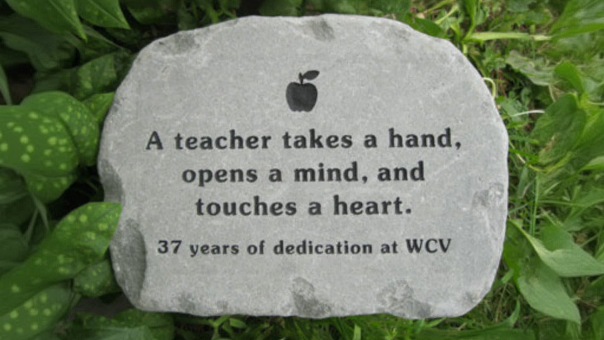 A custom engraved teacher retirement gift garden stone placed outdoors on green grass. The stone features a black engraved apple icon and the quote: 'A teacher takes a hand, opens a mind, and touches a heart.