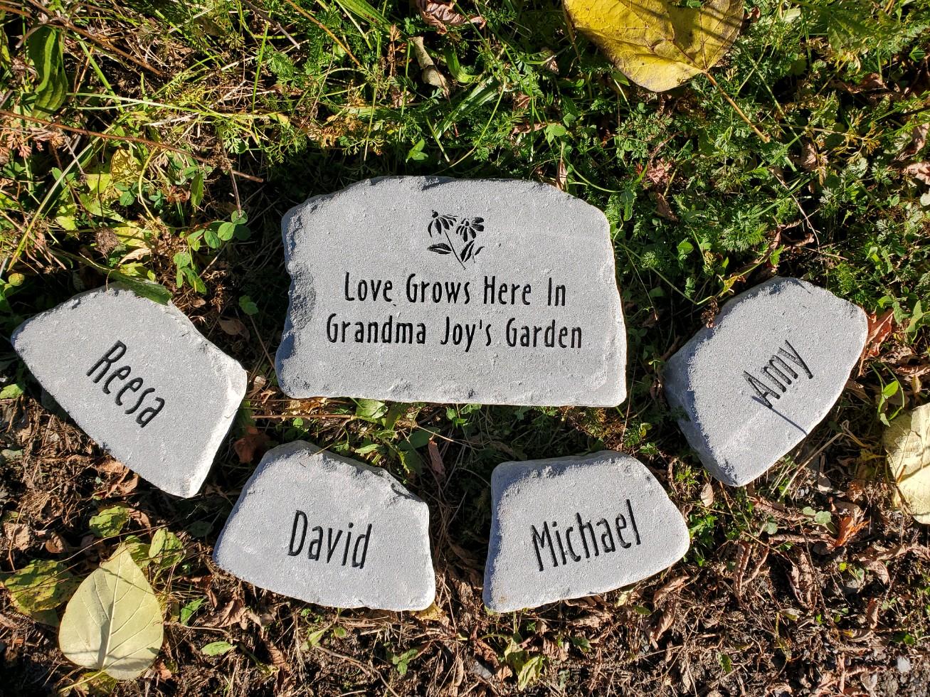 Five personalized garden stones arranged on grass and mulch, with the center stone reading 'Love Grows Here In Grandma Joy's Garden' and the surrounding stones engraved with names including Reeca, David, Michael, and Joy.