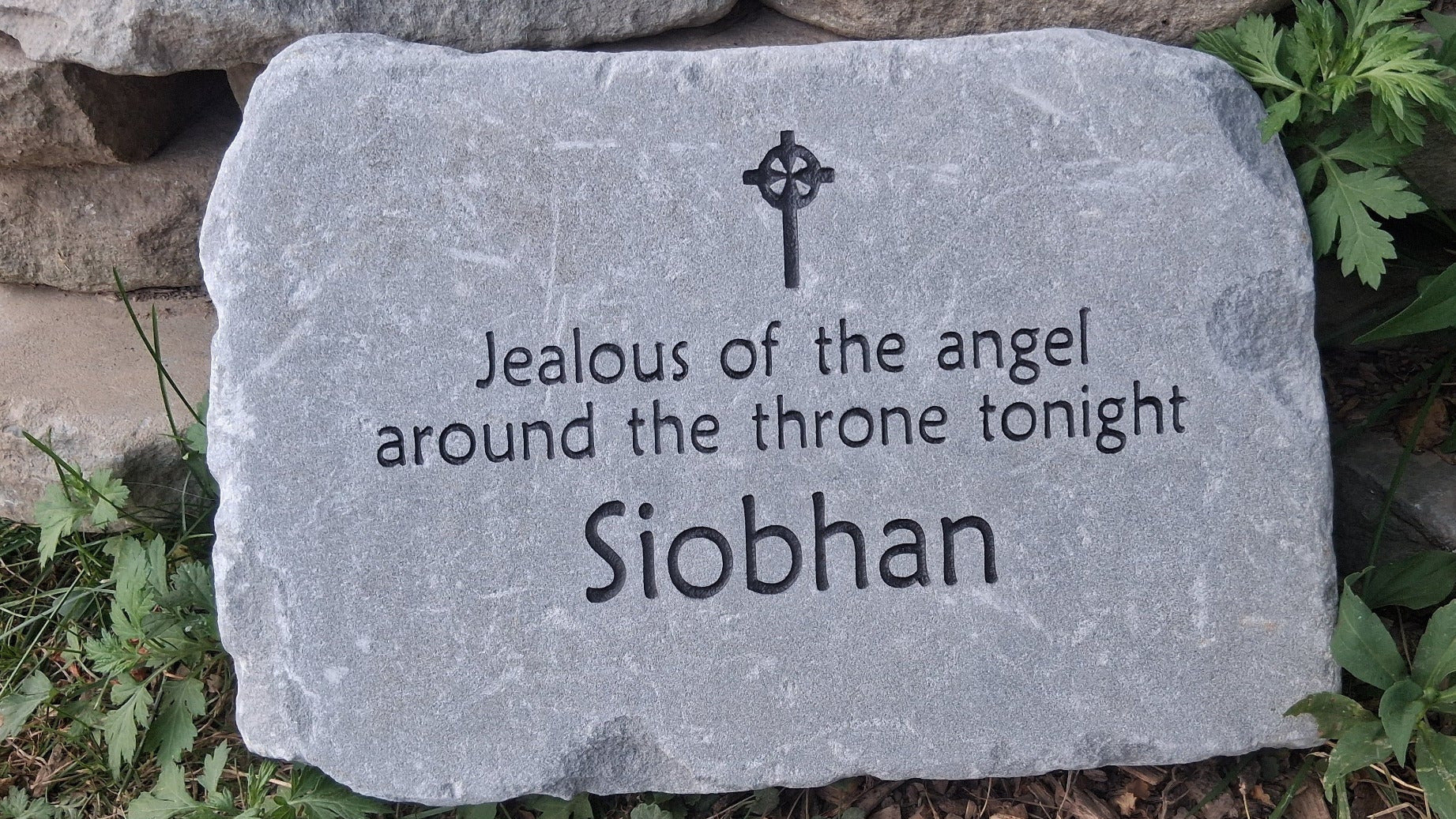 A personalized stone memorial  with a Celtic cross engraving and the text 'Jealous of the angel around the throne tonight Siobhan', placed outdoors on mulch and grass in front of stacked natural stones.