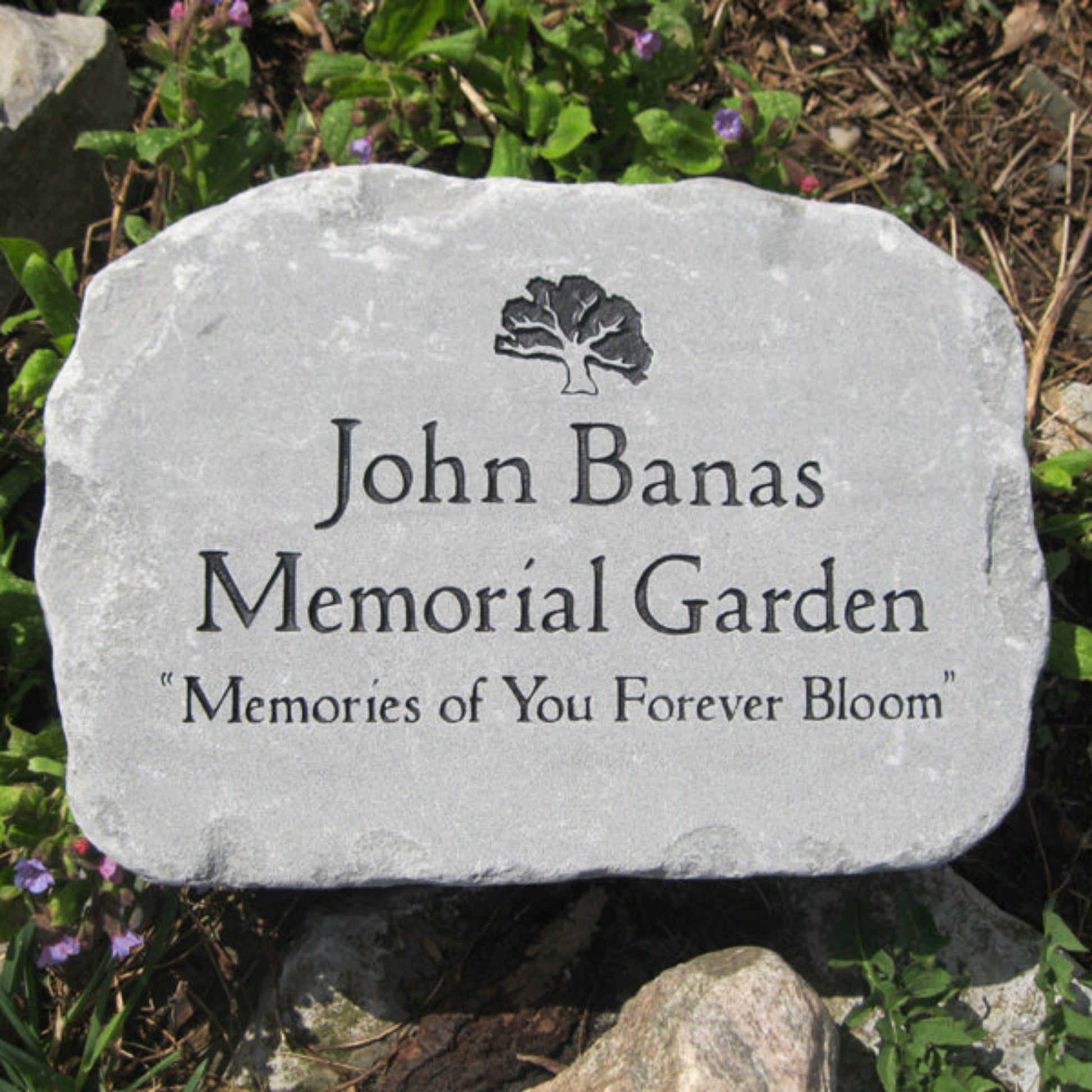 A memorial garden stone plaque reading 'Memorial Garden' with the phrase 'Memories of You Forever Bloom' and a small tree icon, placed outdoors among rocks and greenery.