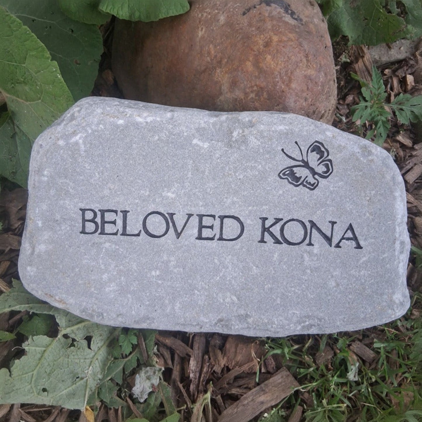 A personalized bluestone garden stone engraved with 'BELOVED KONA' and a butterfly design, placed outdoors on mulch next to green leaves and a brown decorative rock.