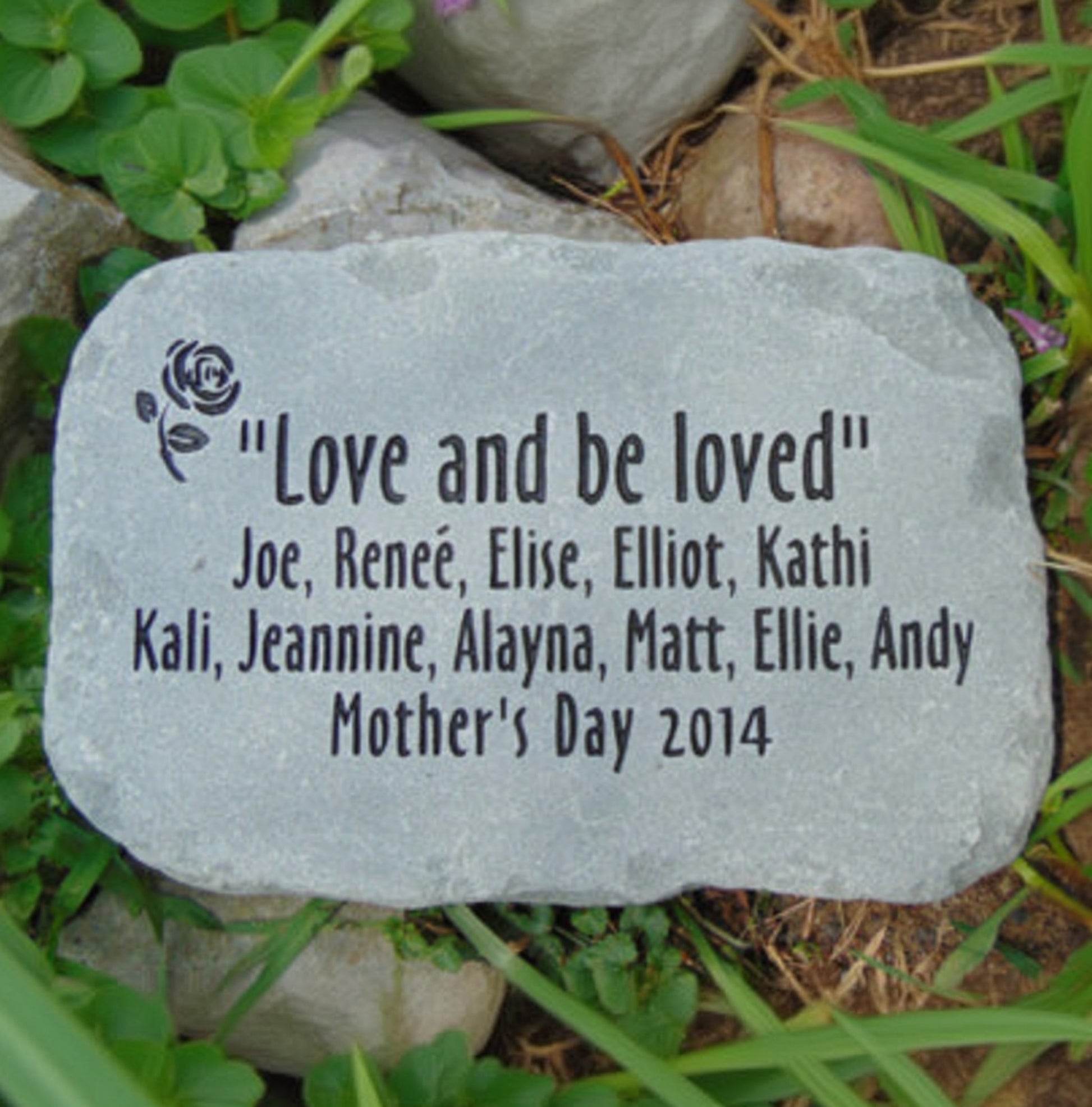 A personalized natural stone with engraved text reading "Love and be loved" and a list of names including Joe, Renée, Elise, Elliot, Kathi, Kali, Jeannine, Alayna, Matt, Ellie, and Andy, dated Mother's Day 2014. The stone is photographed outdoors on grass and soil.