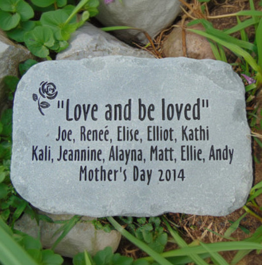 A personalized natural stone with engraved text reading "Love and be loved" and a list of names including Joe, Renée, Elise, Elliot, Kathi, Kali, Jeannine, Alayna, Matt, Ellie, and Andy, dated Mother's Day 2014. The stone is photographed outdoors on grass and soil.