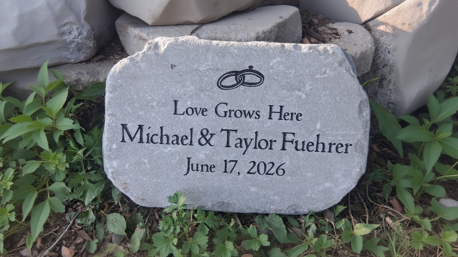 A personalized gray anniversary garden stone with engraved text reading 'Love Grows Here' and  wedding ring graphics placed outdoors among rocks and green foliage