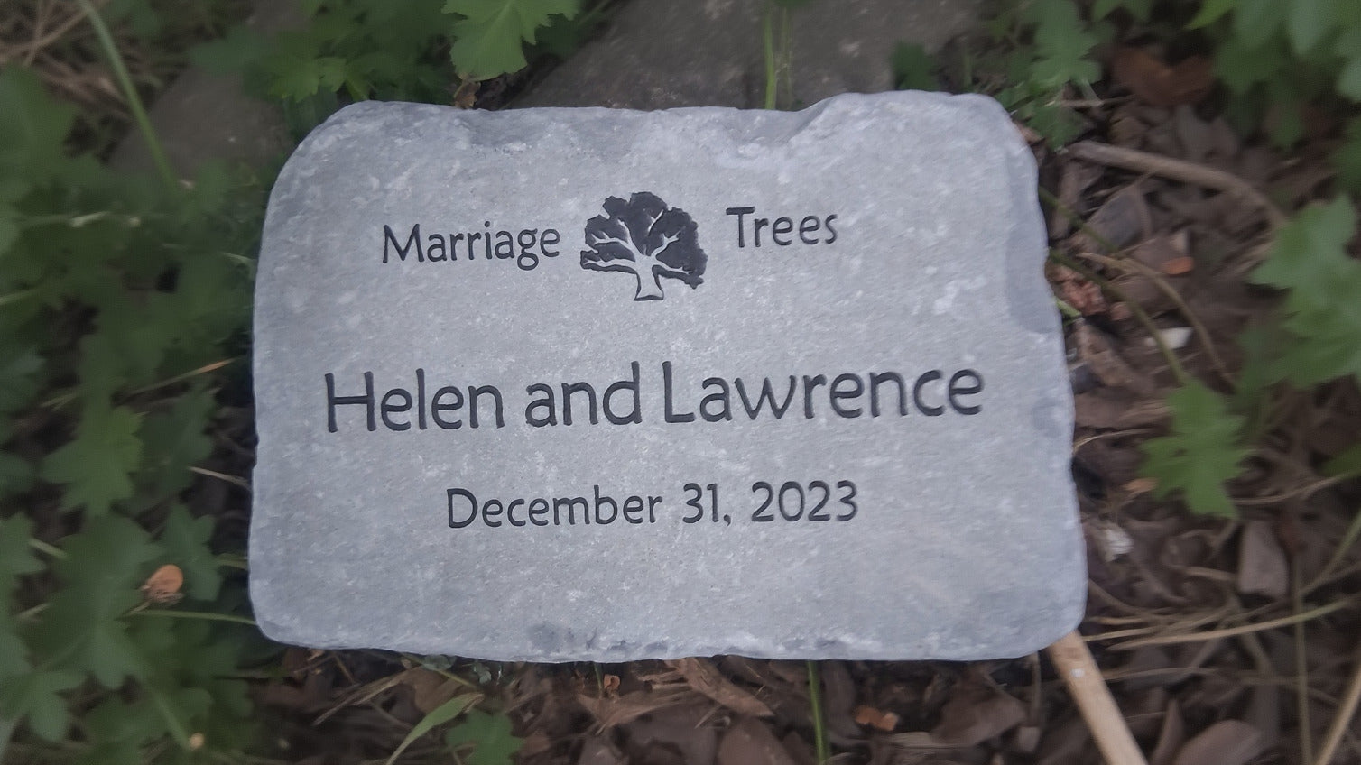 A custom engraved bluestone anniversary gift stone placed outdoors on a bed of mulch and green plants. The stone features the engraved text 'Marriage Trees' with a tree logo.
