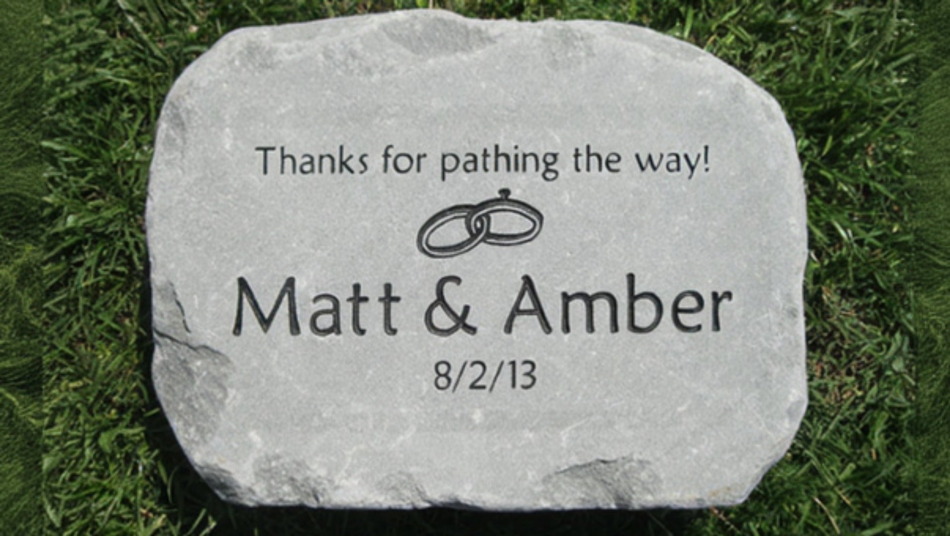 A personalized bluestone plaque engraved with the text 'Thanks for pathing the way! Matt & Amber 8/2/13' and a ring motif, placed on green grass.