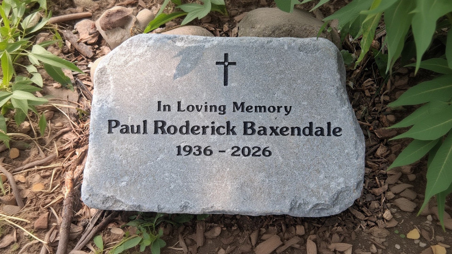 A personalized memorial garden stone with a cross engraving, reading 'In Loving Memory ... ', placed on a bed of mulch and green plants.