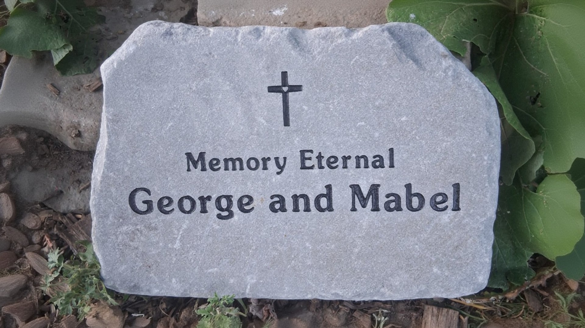 A personalized bluestone garden memorial stone with a black cross and the engraved text 'Memory Eternal George and Mabel', placed outdoors among green leaves and mulch.