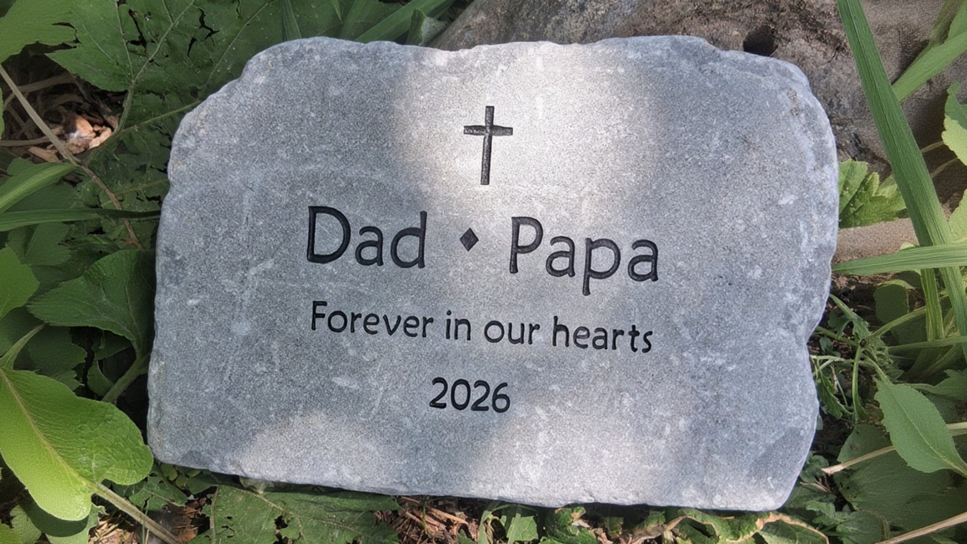 A personalized gray stone memorial stone with a cross, engraved with the text 'Dad • Papa', 'Forever in our hearts', and '2026', placed outdoors on green foliage and soil.