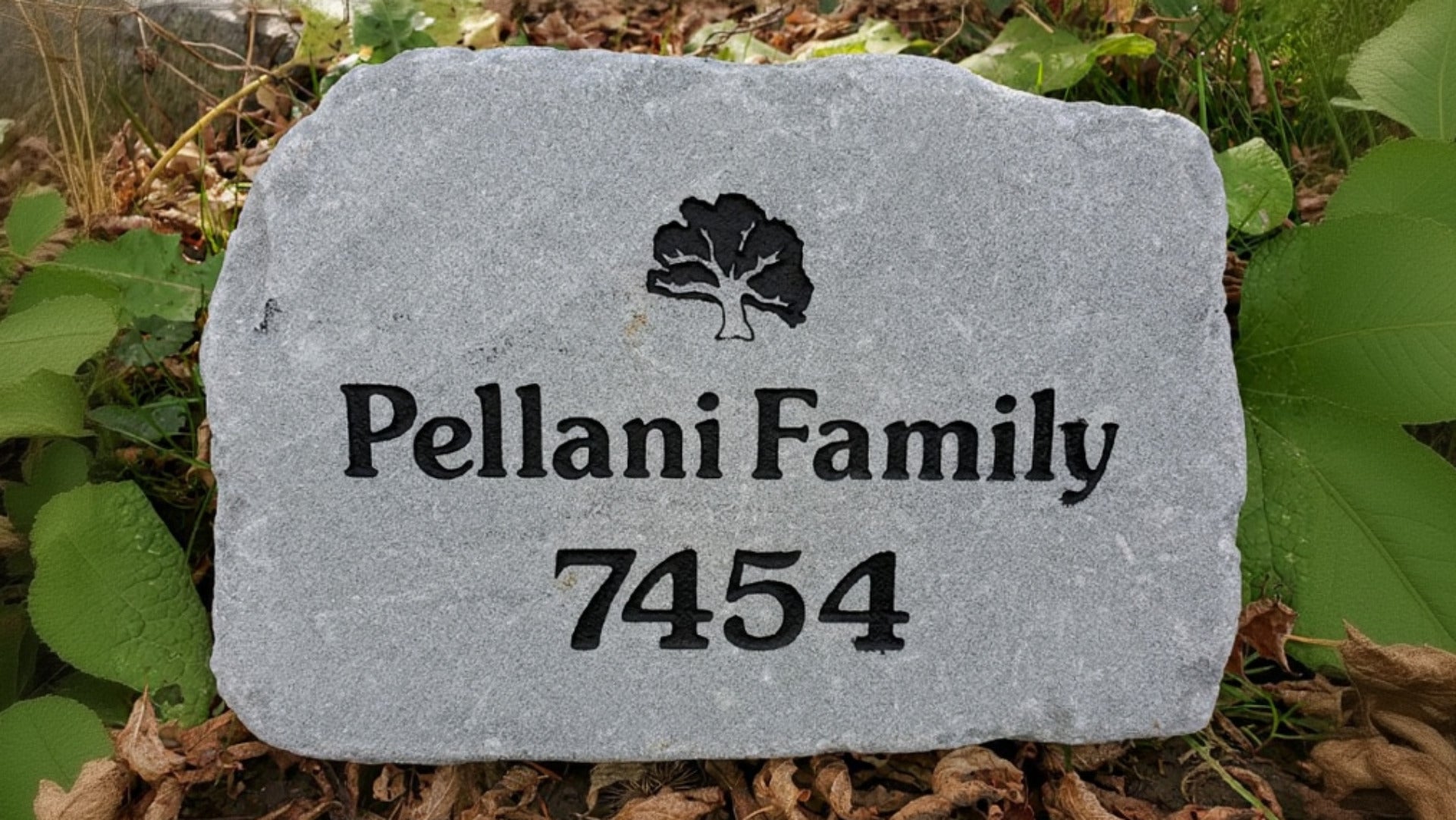 A custom engraved  natural stone address marker with a black tree logo, the text '7454', placed outdoors on dry leaves and green foliage.
