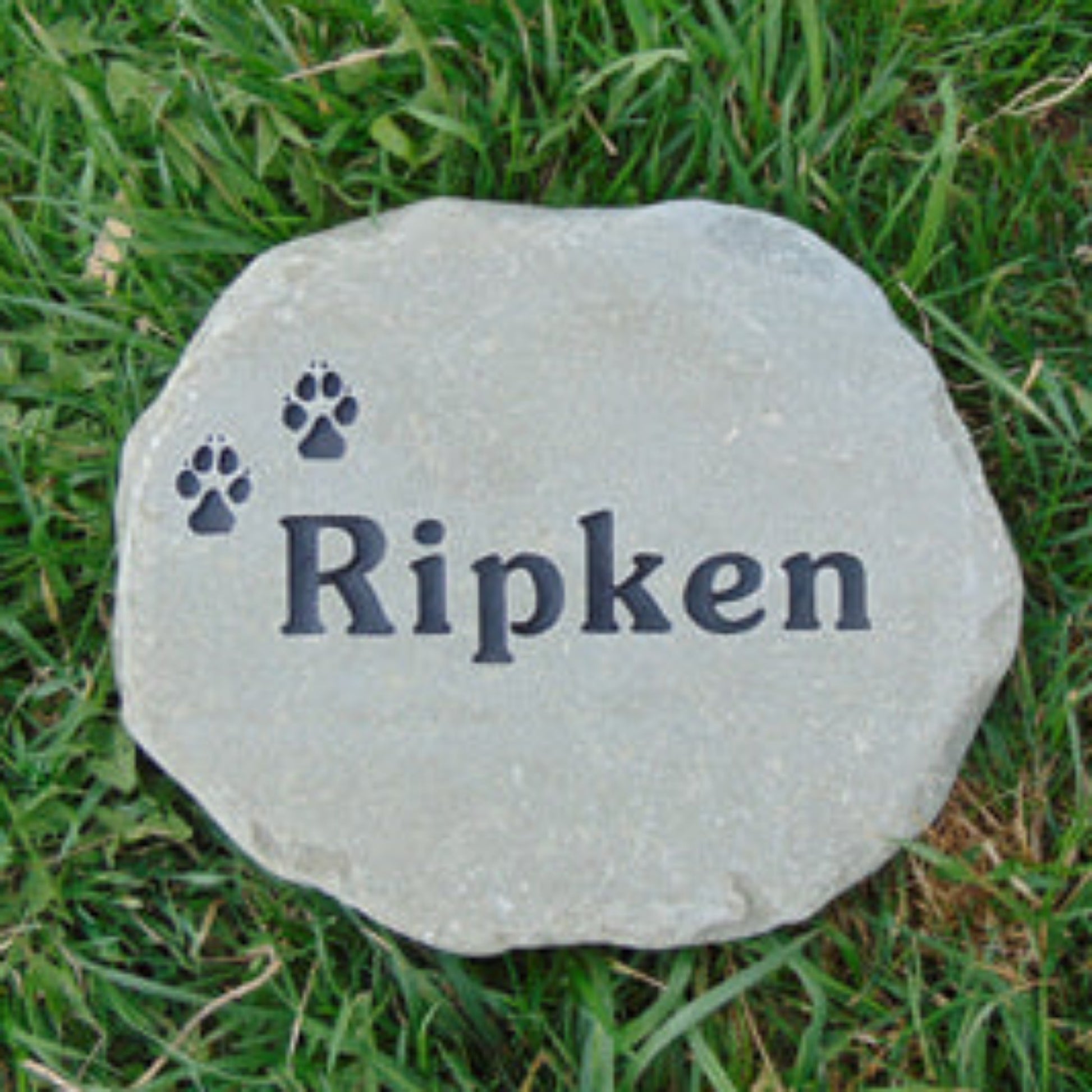 A round gray stone pet memorial marker engraved with the name 'Ripken' and two paw prints, placed on green grass.