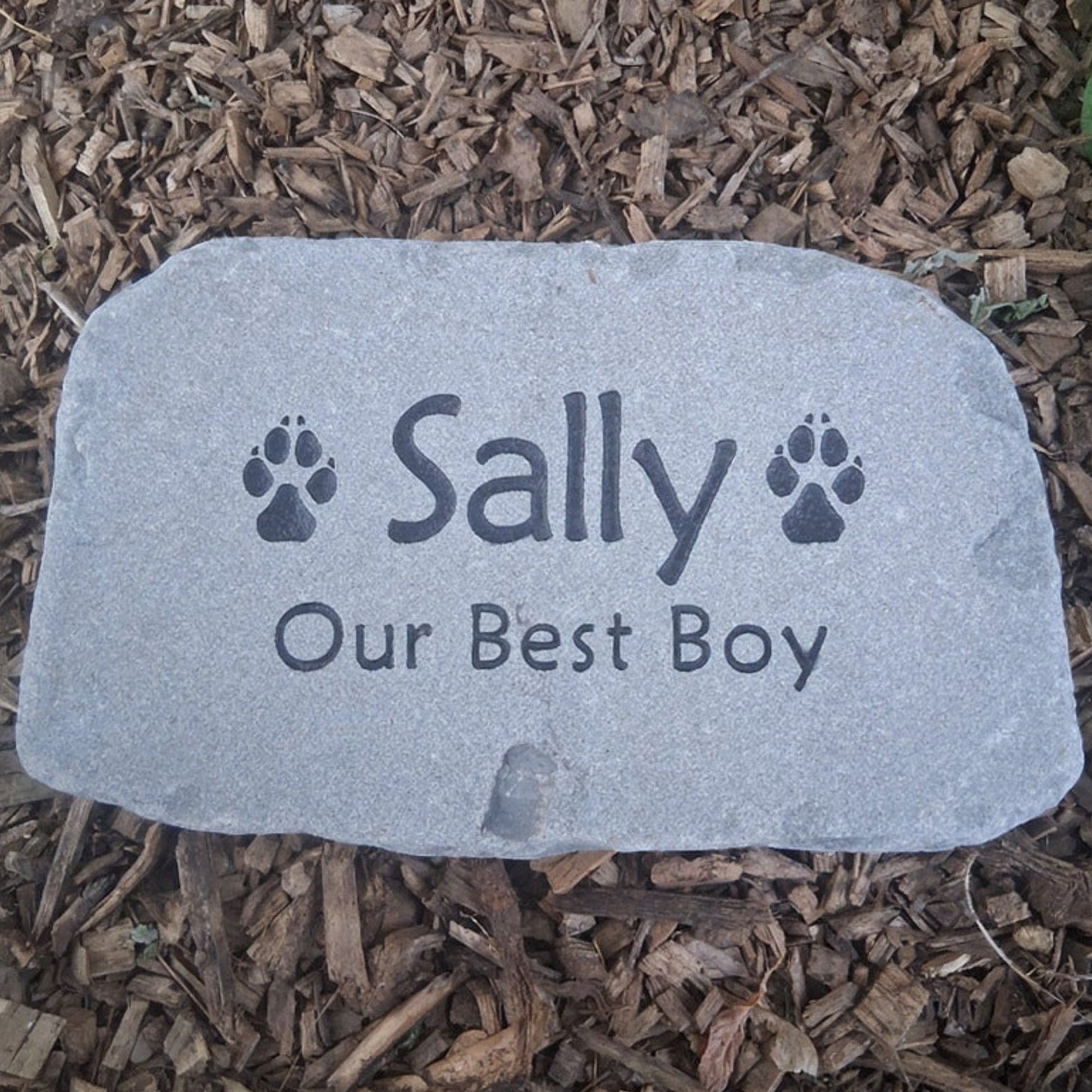 A custom pet memorial garden stone engraved with the name 'Sally', 'Our Best Boy', and paw prints, placed on a bed of brown wood mulch.