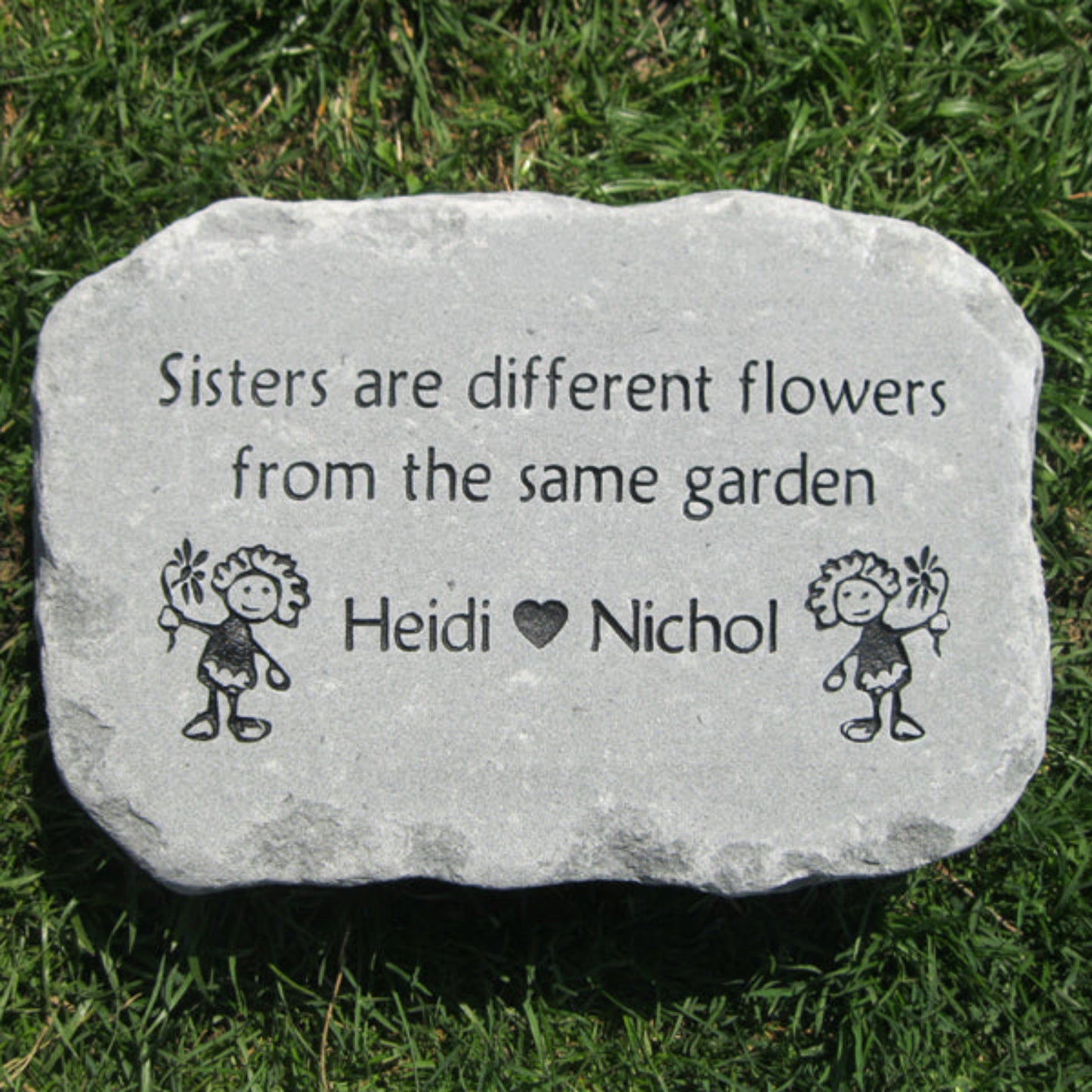 A personalized gray stone garden marker engraved with the quote 'Sisters are different flowers from the same garden', the names 'Heidi' and 'Nichol' with a heart between them, and two small stick-figure drawings, placed on a green grass background.