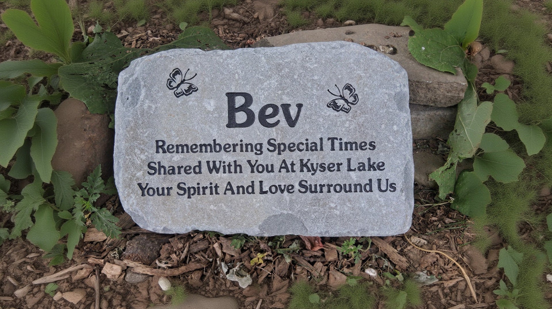 A personalized gray stone memorial garden marker with engraved black text and butterfly designs, placed on a bed of mulch and green leaves.
