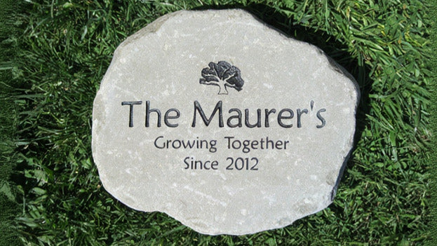 A round natural stone garden marker engraved with 'Growing Together Since 2012' and a small tree logo, placed on green grass.
