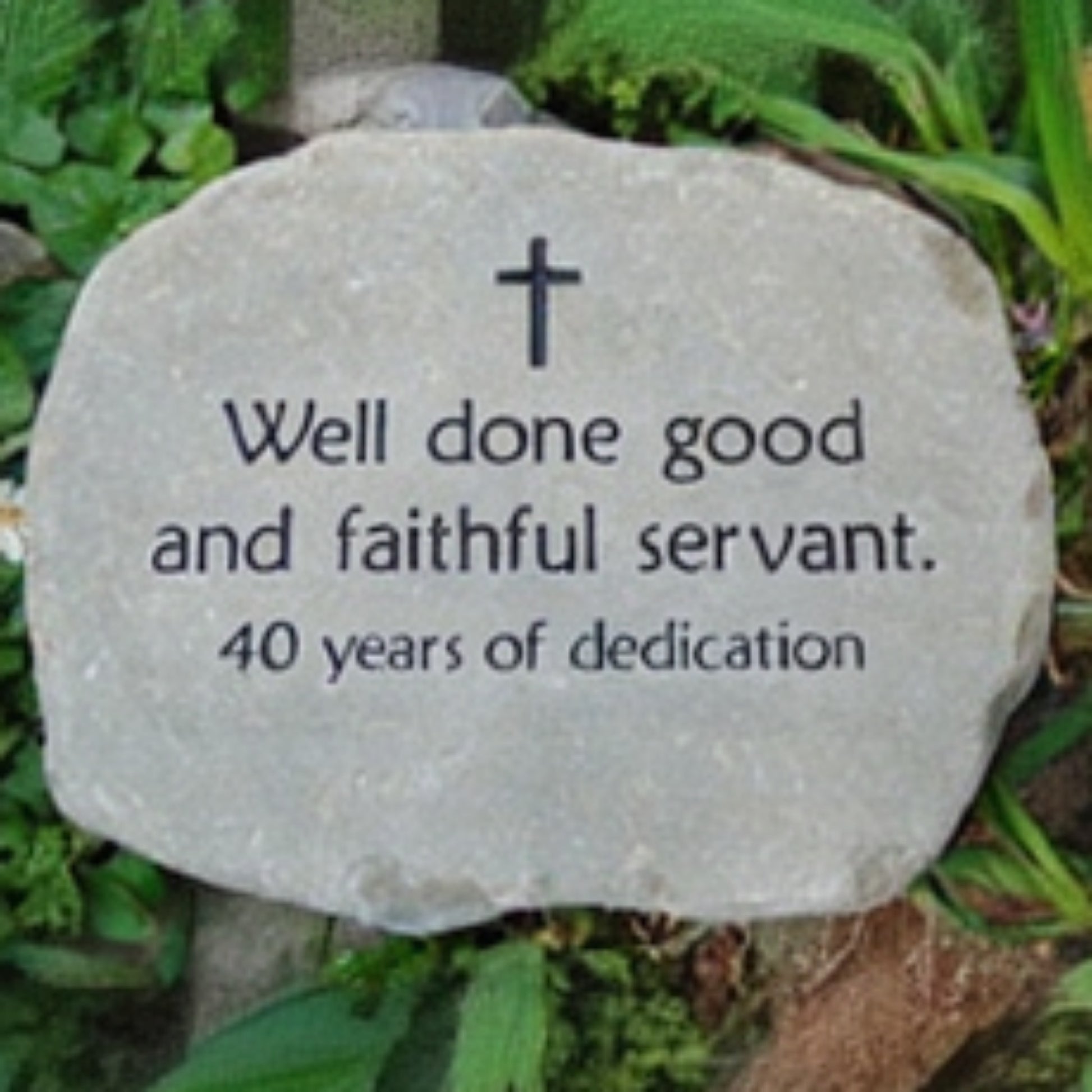 A natural stone plaque with engraved black text reading 'Well done good and faithful servant. 40 years of dedication' and a small cross at the top, placed outdoors among green plants.