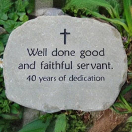 A natural stone plaque with engraved black text reading 'Well done good and faithful servant. 40 years of dedication' and a small cross at the top, placed outdoors among green plants.
